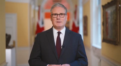 イギリス首相官邸に立つスターマー首相。背後にクリーム色の壁が続き、イギリス国旗が2本立っている