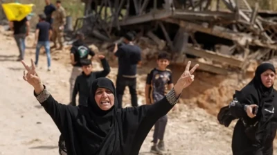 دو زن محجبه با لباس سیاه رنگ، یکی از آنها دستانش را بالا برده وعلامت پیروزی نشان می‌دهد، در پسزمینه ساختمانی ویران و چند کودک دیده می‌شوند