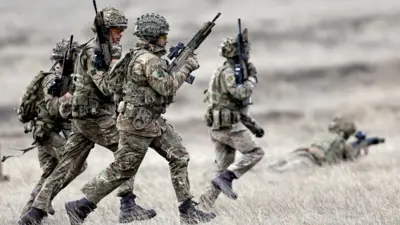 Members of Nato Allied Reaction Force on exercises in Romania. Four soldiers in khaki and helmets carrying automatic weapons run through grassland while another lies prone, apparently about to fire weapon.