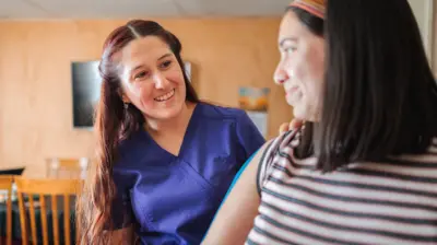 Duas mulheres sorrindo uma para a outra. Uma é médica e está usando uniforme azul, a outra é uma paciente que parece estar sendo examinada.