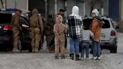 Dört çocuk, arkaları dönük, yalnızca biri kameraya bakıyor. Arka planda iki otomobil ve askeri giyimli kişiler var.