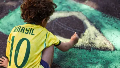 Criança pinta a bandeira do Brasil na rua, parte da decoração para Copa do Mundo