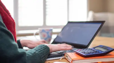 Woman types on laptop computer at kitchen table, with a calculator on top of a notebook