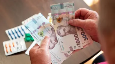 Літня жінка тримає у руках кілька банкнот номіналом 1000 гривень, розкладених віялом. На столі поруч лежать блістери з різнокольоровими пігулками.