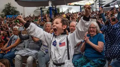 Uma criança fantasiada de astronauta comemora com entusiasmo a aterrissagem da tripulação da Artemis II, enquanto uma mulher agita a bandeira dos EUA durante a transmissão do evento