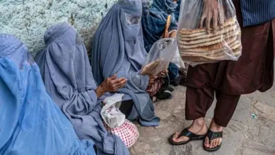 تصویر شماری از زنان افغان که از دست یک مرد نان خشک می‌گیرند. این زنان چادری ابی به تن دارند و در یک صف برای دریافت نان نشسته اند. 