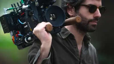 Adolpho Veloso, um homem de barba e óculos escuros, carrega no ombro uma câmera profissional de cinema, equipada com lente grande e diversos cabos e acessórios. Ele veste uma camisa verde-oliva de manga longa e segura a câmera com firmeza pela estrutura lateral, apoiando-a sobre o ombro direito. O fundo é desfocado, com tons de verde que sugerem um ambiente externo, possivelmente em meio à natureza.