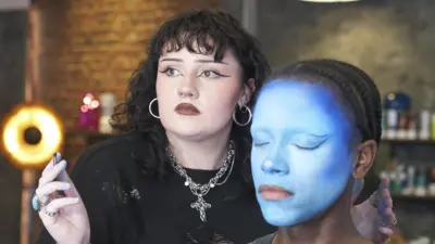 Ailish is standing next to a seated make-up model. Ailish is holding a thin brush in one hand and has two turquoise rings on her fingers. She is looking off to the side, probably at a mirror, rather than directly at the model and has a look of concentration on her face. Ailish has black wavy hair with a fringe and bold black eyeliner. She is also wearing a black distressed jumper, silver hoop earrings and a silver necklace with a cross on it. She also has three silver nose piercings, including a nose ring, stud and septum barbell. The model has her eyes closed and her entire face has been painted using different shades of blue. It is clear that the make-up look has not been complete yet and the model's lips are bare. The model's black hair has been plaited back into cornrows. The background is out of focus but Ailish and the model are in a make-up studio with shelves or products and a ring light behind them. 