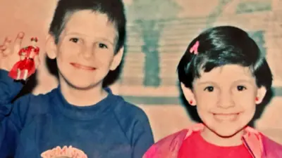 A boy in a blue top and a girl in a pink top, both with dark hair, smile at the camera - the boy is holding up a toy figurine 