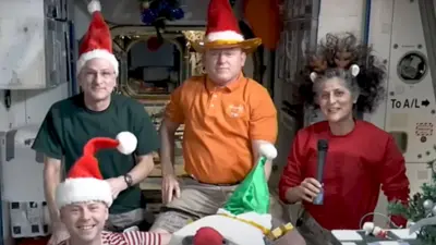 Four astronauts on the International Space Station wearing a mix of Santa hats and reindeer antlers gather together in front of a camera to wish people a merry Christmas