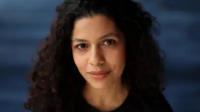 Arianna de Souza-García mira a la cámara. Es una mujer de cabello rizado y oscuro, con camiseta negra, situada frente a un fondo azul oscuro.