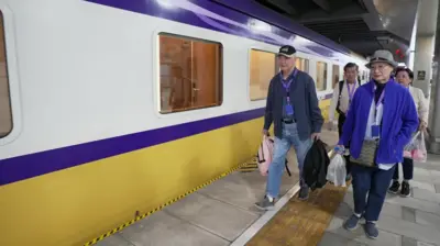 A group of four people - two men and two women - carrying bags in their hands, the front two wearing hats and blue tops, walk along the platform of a train with stripes of blue, white and yellow
