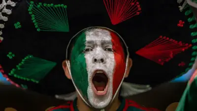 Un hombre con la boca abierta, seguramente está gritando o cantando. Tiene la cara pintada con los colores verde, blanco y rojo, que conforman la bandera de México. Lleva puesto un sombrero de mariachi.