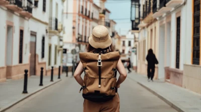 Turystka stoi na środku cichej ulicy w zabytkowym mieście, tyłem do obiektywu. Na głowie ma słomiany kapelusz, a na plecach - brązowy płócienny plecak. Po obu stronach widoczne rzędy domów o kamiennych podmurówkach. Domy mają tynkowane ściany w pastelowych kolorach: jasnożółte, bladoróżowe i białe. Ich drzwi wejściowe i okna obmalowano na żółto lub pomarańczowo. Na dalszym planie widać inną osobę, w czarnej bluzie i spodniach, idącą w dół ulicy. 