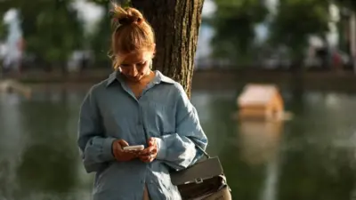 Женщина стоит под деревом и смотрит в телефон. На заднем плане водоем с домиком для уток