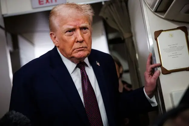 Trump is on Air Force One, wearing a navy ⁣blue suit, a⁤ white ⁤shirt, and a ‌burgundy tie with navy blue dots. ⁣On his suit jacket he wears an American flag⁣ pin.