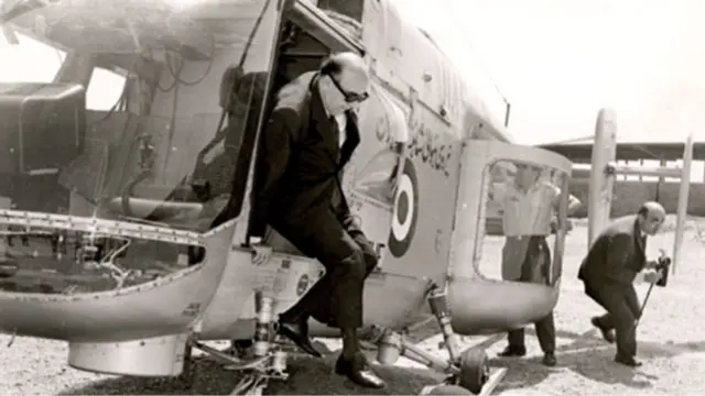 هویدا در حال خروج از هلی کوپتر نظامی؛ شخص دیگری عصا و پیپ او را می آورد