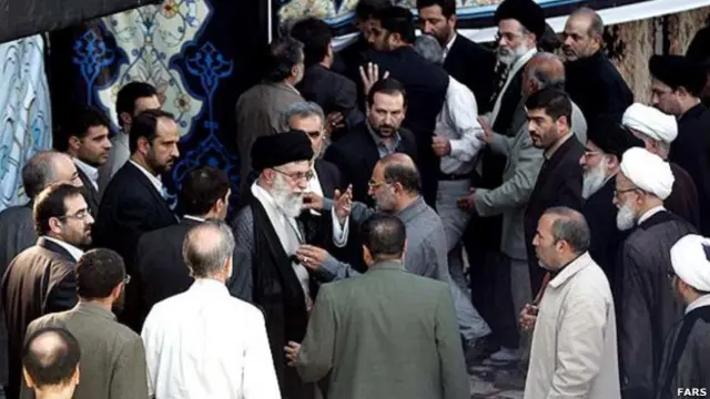 حسین جباری در حال برداشتن چفیه آیتالله خامنهای از دوش او. مصطفی خامنهای هم در پس زمینه عکس دیده میشود