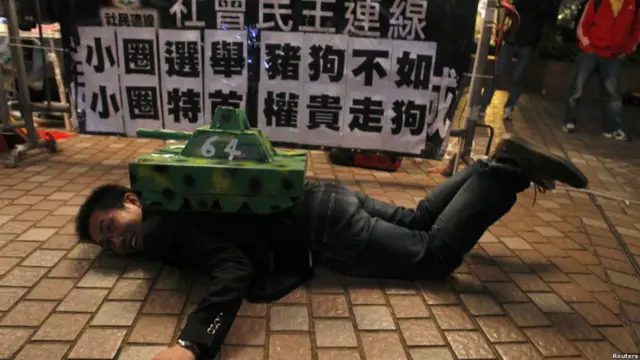 一名示威者趴在印有64标志的坦克模型下。