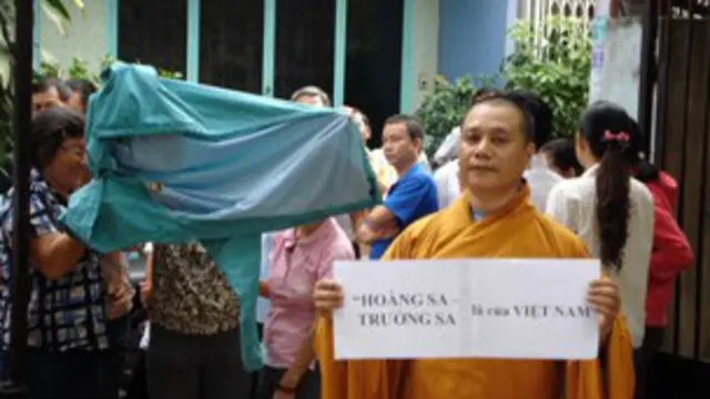 Đại đức Thích Viên Hỷ cầm biển "Hoàng sa – Trường sa là của Việt Nam" ở chùa Giác Hoa, Tp Hồ Chí Minh trong khi có người lấy áo mưa che mặt vì sợ bị nhận diện. Ảnh: PTTPGQT