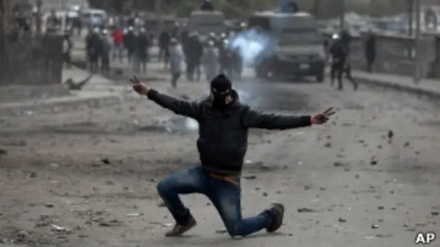 Individuo enmascarado hace el signo de la victoria en mitad de unos disturbios