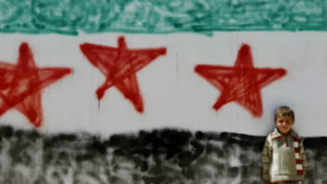 کودکی در کنار دیواری با نقش پرچم سوریه بر آن