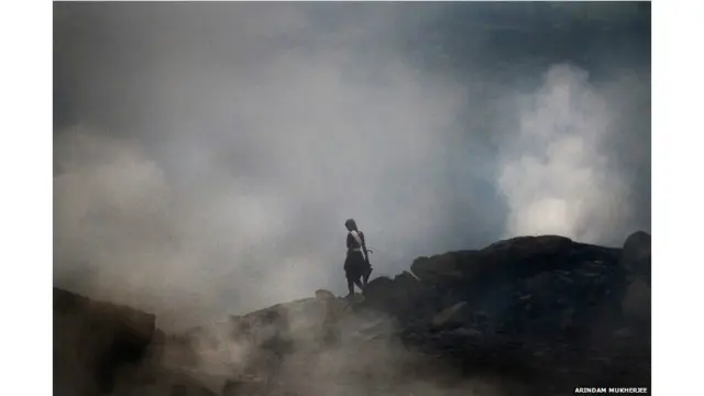 तस्वीर में एक युवती ज़मीन में लगी आग और उससे निकलनेवाली ज़हरीली गैस के बीच गुज़रती हुई. जलती खदानों के आसपास का तापमान इतना ज़्यादा होता है और हवा इतनी ज़्यादा प्रदूषित होती है कि यहां के बाशिंदों को स्वास्थ्य संबंधी तमाम परेशानियां पैदा हो गई हैं.