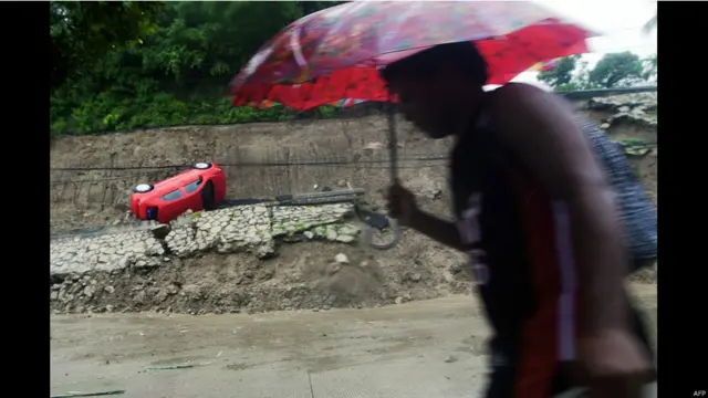 Un hombre con paraguas pasa juntoa un conche volcado por la fuerza de las aguas. 