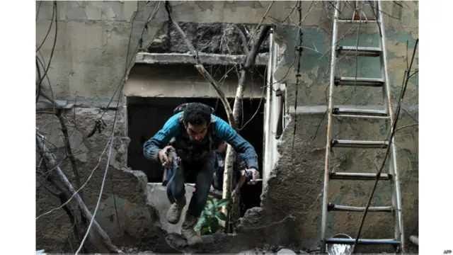 شام میں ایک باغی کلاشنکوف اٹھائے نشانچیوں سے بچتے ہوئے ایک شکستہ دیوار سے نکل کر بھاگ رہا ہے۔