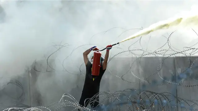 ایک شخص دیوار کے اوپر سے سکیورٹی اہلکاروں پر آگ بجھانے والے آلے سے گیس پھینک رہا ہے۔ 