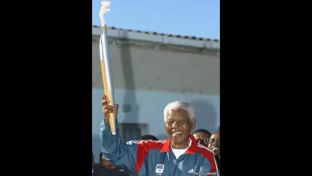 نیلسن منڈیلا 2004 میں اولمپک ٹارچ کو اٹھا کر روبن جزیرے کی اس جیل میں کھڑے ہیں جہاں انھیں 17 برس تک قید رکھا گیا۔ 