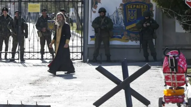 أحد رجال الدين يمر أمام جنود يعتقد أنهم ينتمون للجيش الروسي (يمين) في قرية بيريفالنوي في القرم، بينما يقف أفراد القوات الأوكرانية خلف البوابة.