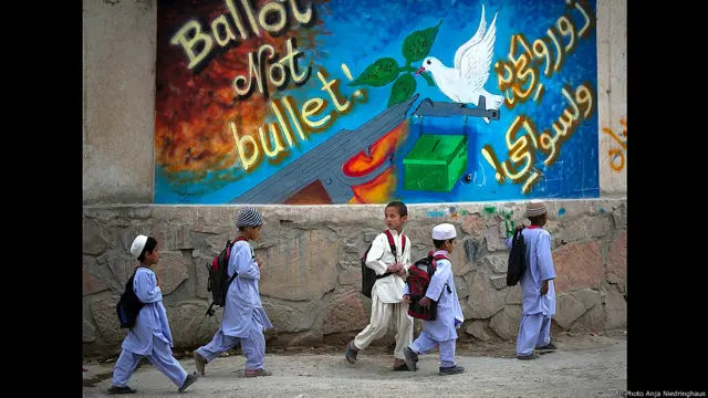 Niños con mochilas en sus espaldas pasan junto a una pancarta electoral 