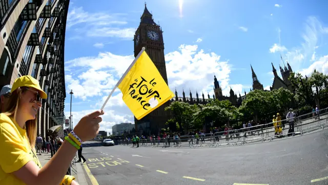数万人聚集在沿途各地观看这一比赛。一名围观者在伦敦议会大楼前挥舞环法自行车大赛旗帜。