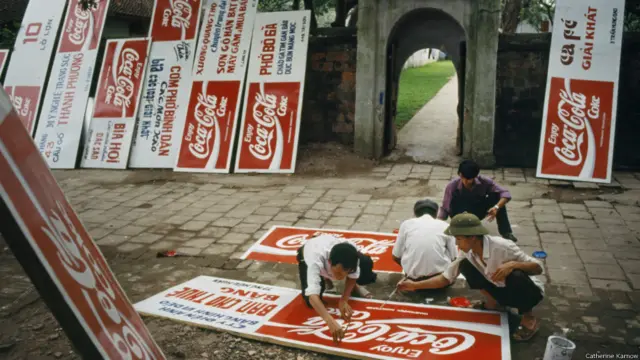 COCA COLA TỚI VIỆT NAM, 1994. Như dấu hiệu báo trước cho cuộc cách mạng tiêu dùng, các công nhân sơn bảng quảng cáo Coca Cola. Việc áp dụng những chính sách Đổi mới của chính phủ khuyến khích đầu tư nước ngoài đã có hiệu quả rõ rệt. Cùng với John Deere và Hewlett Packard là Coca Cola, một sản phẩm biểu tượng của nước Mỹ. 