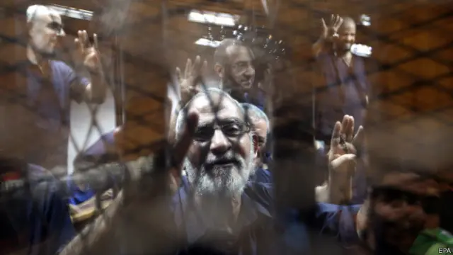 Мухаммед Бади на процессе, здороваясь со сторонниками, сложил пальцы в специальный жест, которым противники Сиси приветствуют друг друга с конца 2013 года