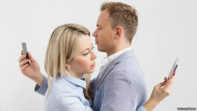 Una pareja se abraza y mira sus respectivos teléfonos a espalda de la otra