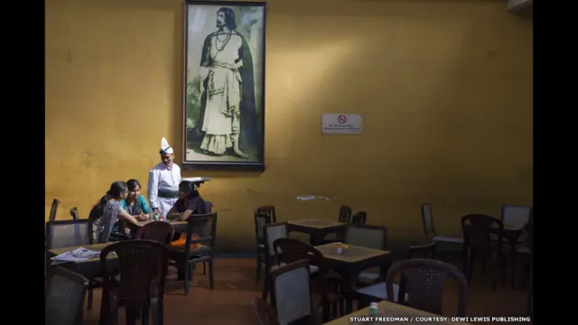 Hoton wani ma'aikacin shagon shan shayi da ke Kolkata, yana bai wa yara 'yan makaranta shayi, daga saman bango ana iya kallon zanen hoton Rabindranath Tagore.