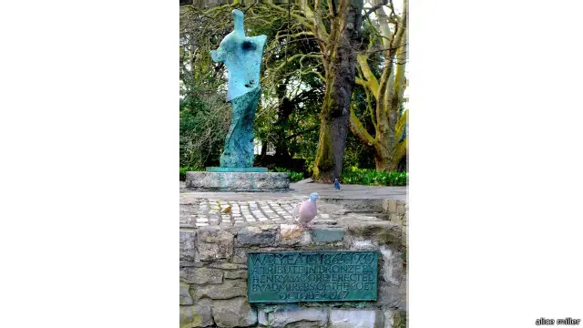Patung Yeats karya Henry Moore tertutup bayangan pohon serta angsa-angsa jinak yang berenang di aliran sungainya.