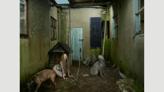 Oxana Malaya, Ukraine, 1991. Những bức ảnh trong của Julia Fullerton-Batten kể các câu chuyện người thật, việc thật. Đây là Oxana Malaya người Ukraine. Theo Fullterton-Batten, Oxana được tìm thấy khi đang sống trong chuồng chó hồi 1991. Khi đó cô bé tám tuổi và đã sống với chó được sáu năm. Cha mẹ cô nghiện rượu. Một đêm nọ, họ đã đã bỏ cô bé ở ngoài nhà. Trời lạnh, em bé ba tuổi bò về phía chuồng chó ở nông trại. Cô bé đã được những chú chó lai trong chuồng ủ ấm, và có lẽ nhờ vậy mà bé đã không chết cóng. Oxana chạy bằng bốn chi, thè lưỡi, nhe răng và sủa. Nay Oxana sống trong một phòng mạch tại Odessa (Ukraine), cô giúp chăm sóc gia súc, gia cầm trong nông trại của bệnh viện.