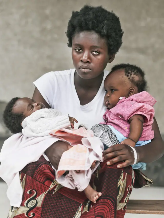 A los 9 años, Marie Asafiwe fue testigo del asesinato de su padre y hermanos, y de la violación de su madre, producto de la cual nació un niño. Un año después mataron delante suyo a su madre y hermano. Cuando fue retratada, tenía 17 años y un hijo que, por ser fruto de una violación, no aceptaba. En la imagen también carga a una niña huérfana que estaba amamantando. Meses después Sartorotti supo que Marie había aceptado a su hijo, a quien bautizó Armando en su honor.