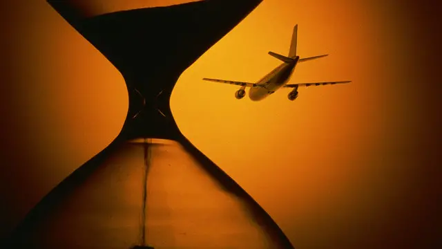 Un avión en se aleja por el horizonte; un reloj de arena en primer plano