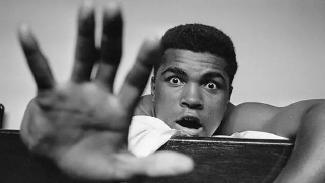 تصویری از کلی روی تخت اتاق هتلش در لندن، در حالی که با دستش نشان میدهد در چند راند هنری کوپر را شکست میدهد، می ۱۹۶۳