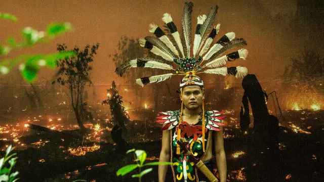 Seorang berpakaian adat dengan latar belakang foto lahan gambut yang terbakar. Kebakaran lahan gambut yang tak terkendali bisa meluas berkilo-kilometer di bawah permukaan tanah mengakibatkan kabut asap yang mematikan.