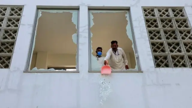 হাসপাতালের ভাঙা দুটি জানালা দিয়ে বাইরে কাচের গুঁড়ো ফেলছেন দুই জন কর্মী
