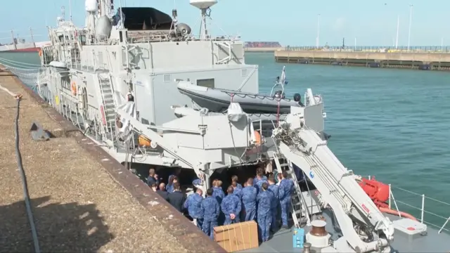 Szary okręt wojenny z dźwigiem na rufie zacumowany w porcie. Na pokładzie znajduje się kilku członków załogi w mundurach marynarki wojennej.