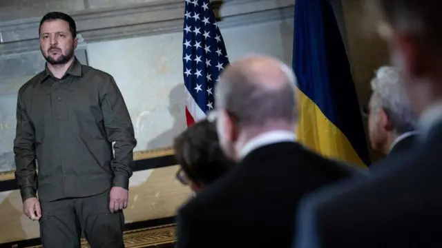 Volodymyr Zelensky em pé, com olhar sério e ao lado de bandeira americana, observado por plateia