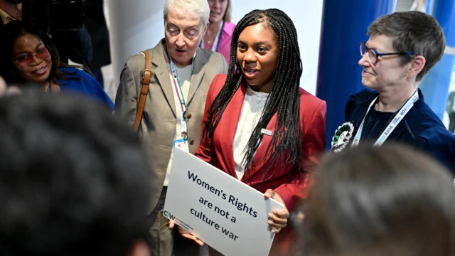 Kemi Badenoch na conferência conservadorabwin withdrawal2024, segurando um cartaz com os dizeres "Os direitos das mulheres não são uma guerra cultural"