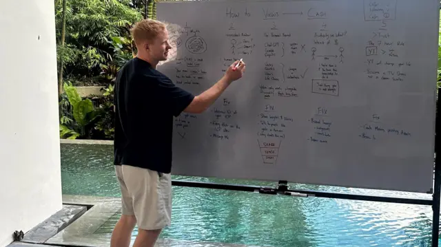 Sol, vestindo uma camiseta preta e shorts bege, escreve em um quadro branco em frente a uma piscina. 