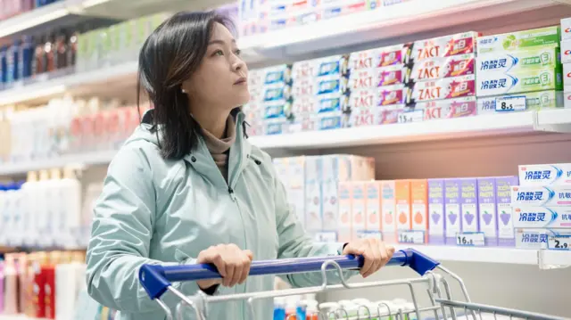 Mulher empurra carrinho de supermercado em frente a uma gôndola com diferentes marcas de creme dental